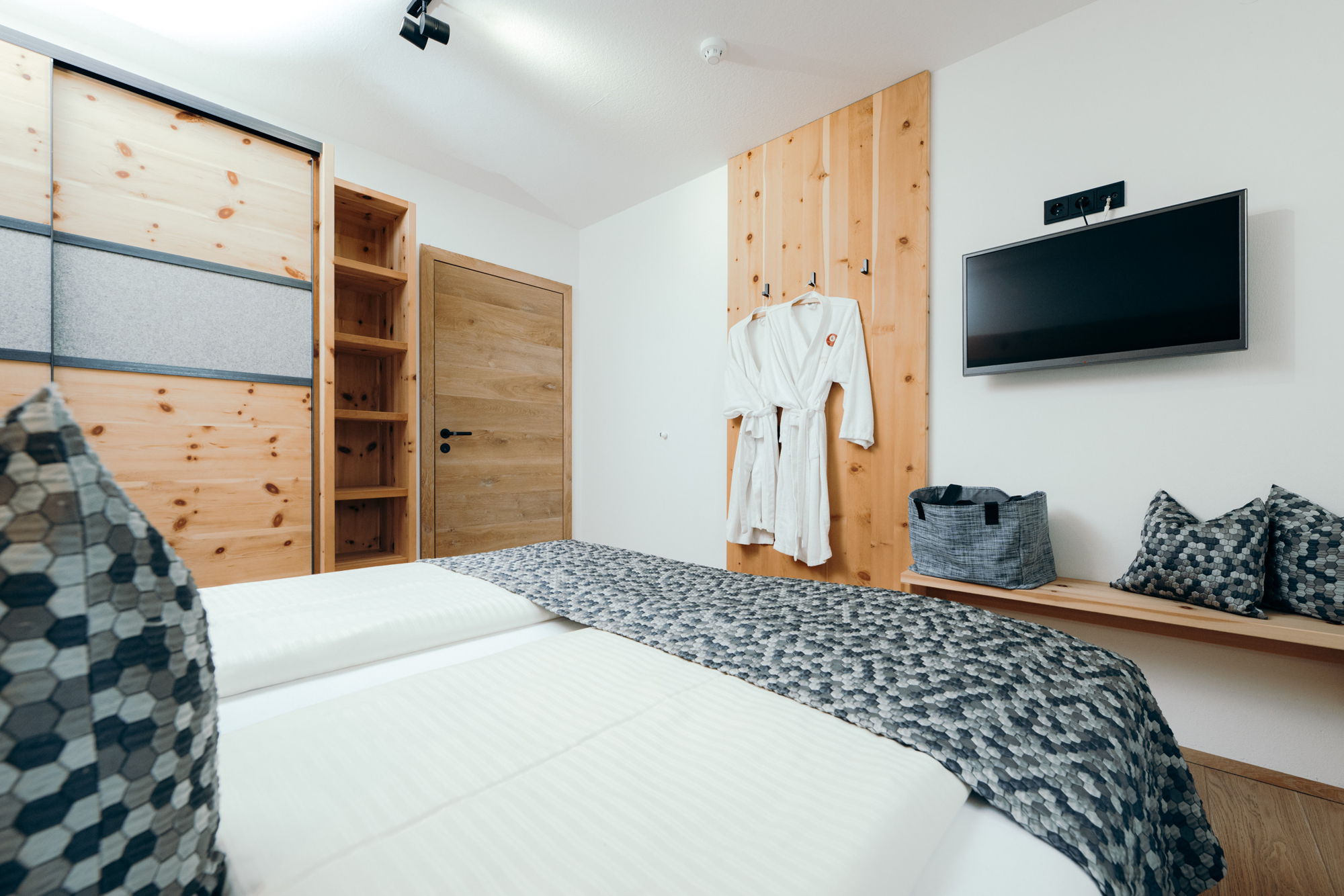 Ein weiß bezogenes Bett mit mehrfarbig-grauem Überzug, im Hintergrund ein Fernseher und weiße Bademäntel.
