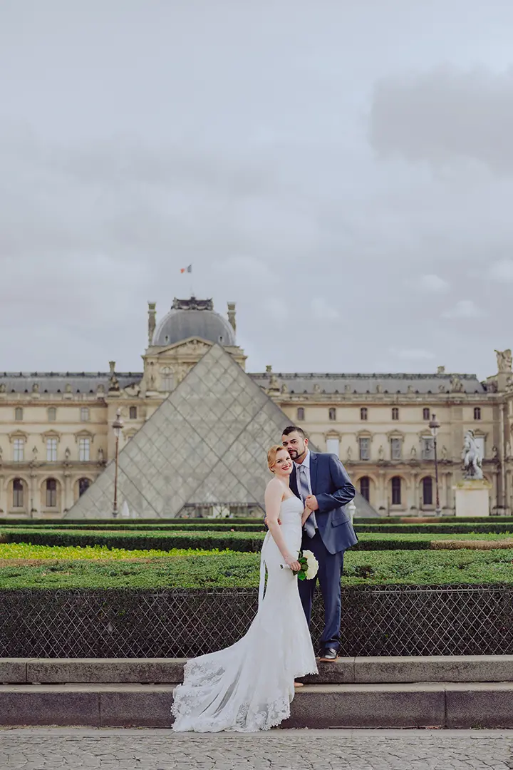 Une belle mariée et un beau marié posent pour des photos de mariage devant le musée du Louvre, avec des haies vertes derrière eux et la célèbre pyramide visible par temps nuageux.