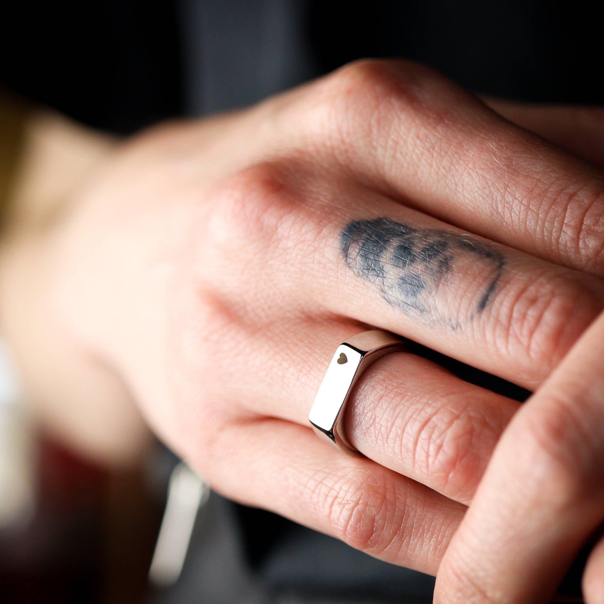 A close up image of a woman's right hand with a skull tattoo on the middle finger and a stainless steel signet ring on the third finger.