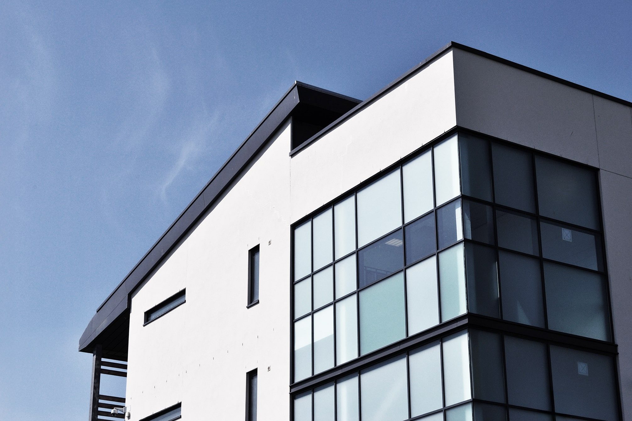 Modern white building with large glass windows and sharp geometric lines, set against a clear blue sky. The photo captures the upper corner and roof of the structure.