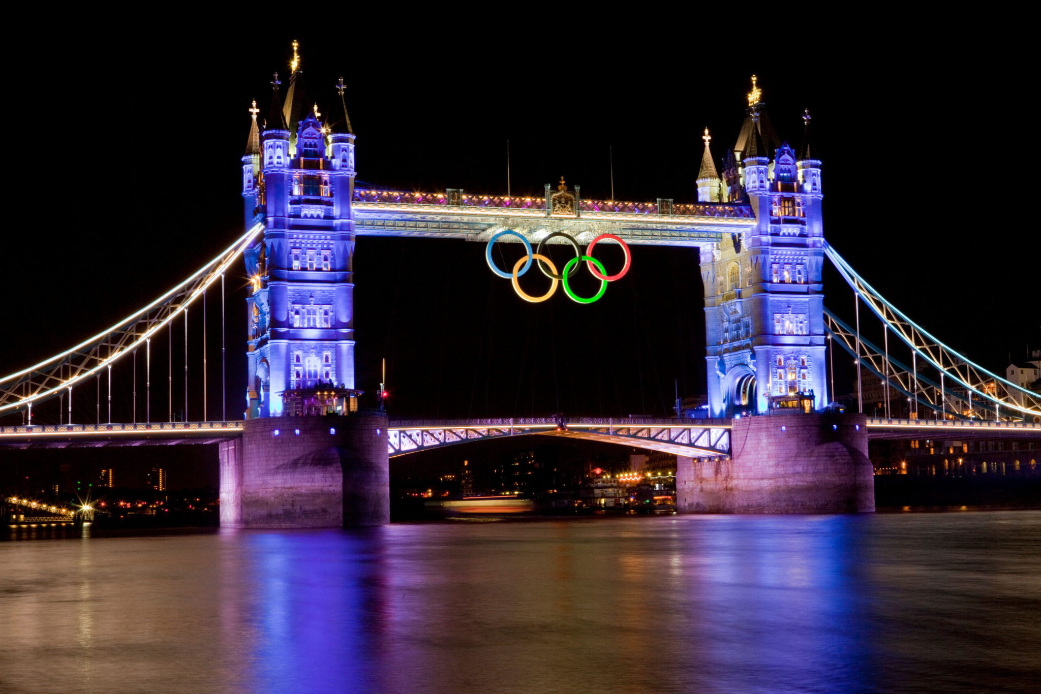 Tower Bridge in London illuminated at night, with large Olympic rings hanging between its towers over the River Thames. The reflection of the lights and rings is visible on the water below.