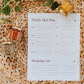 A weekly meal plan sheet lays ready to be filled out on top of a wicker backdrop with spice jars laying on it