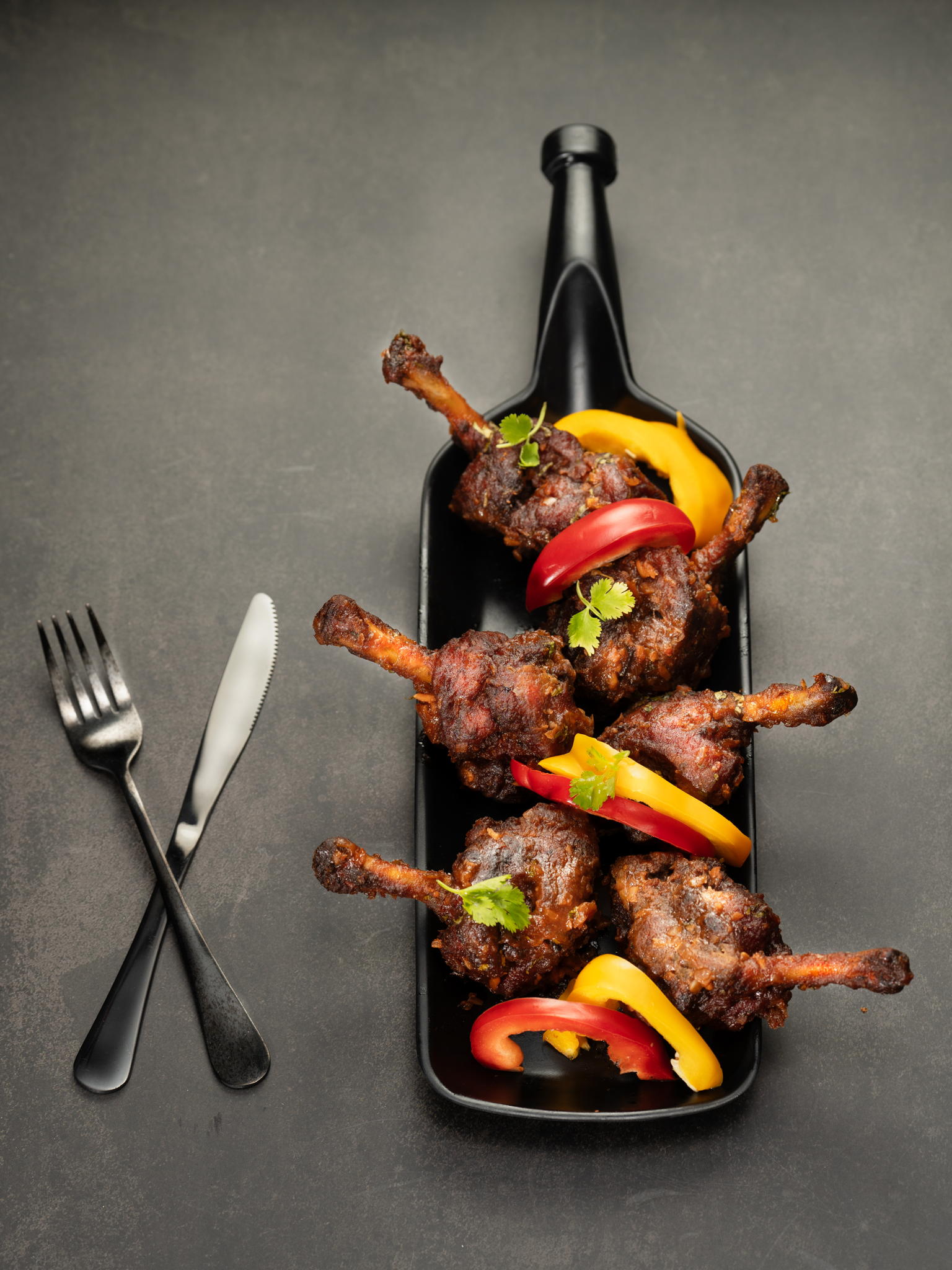 Skewers of chicken lollipops and colorful bell peppers on a metal tray with a red napkin, knife, and fork
