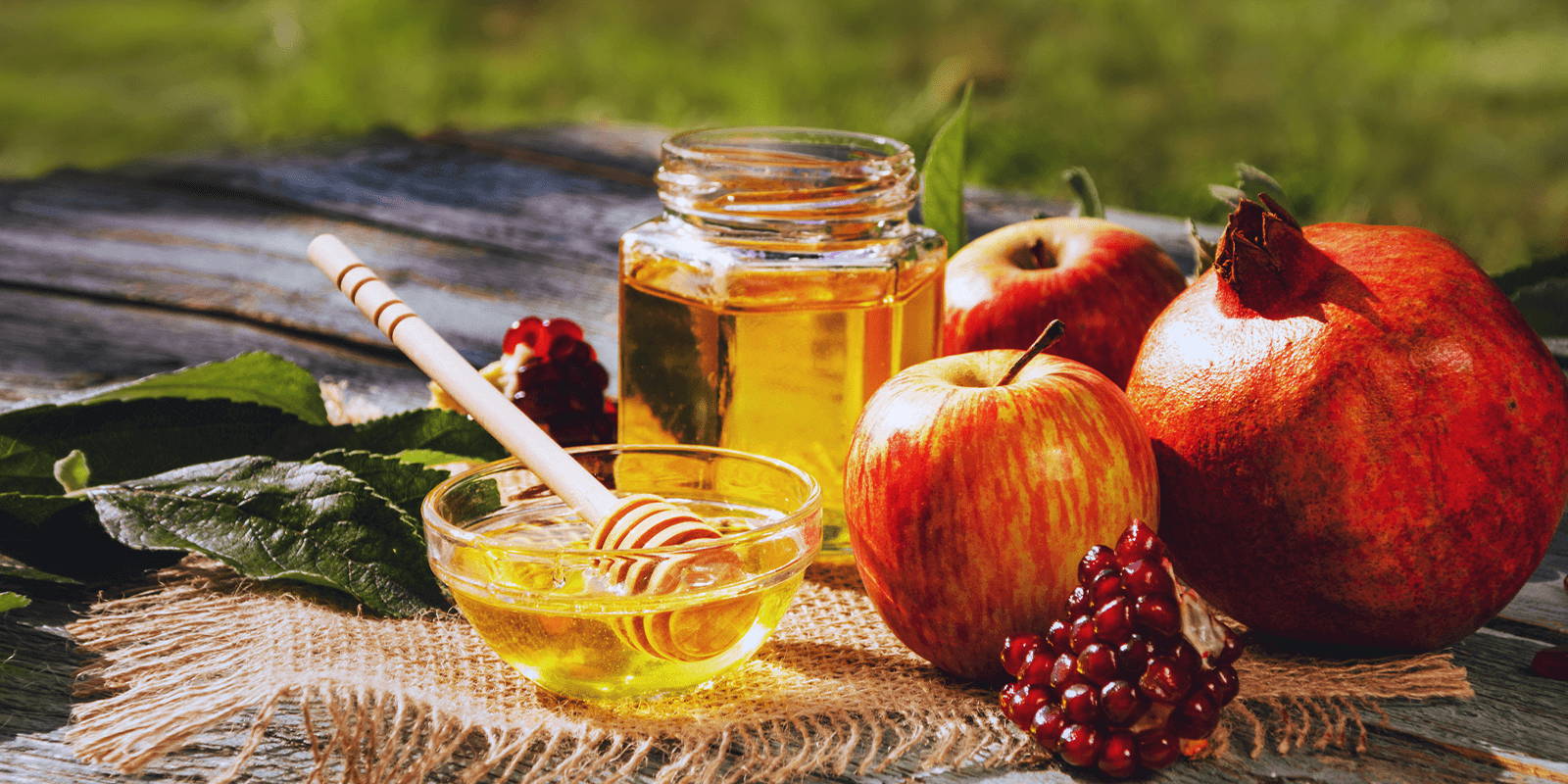 A small glass bowl of honey with a wooden dipper, next to a jar of honey, two apples, leaves, a whole pomegranate, and pomegranate seeds, atop a burlap cloth on a wooden table outdoors.