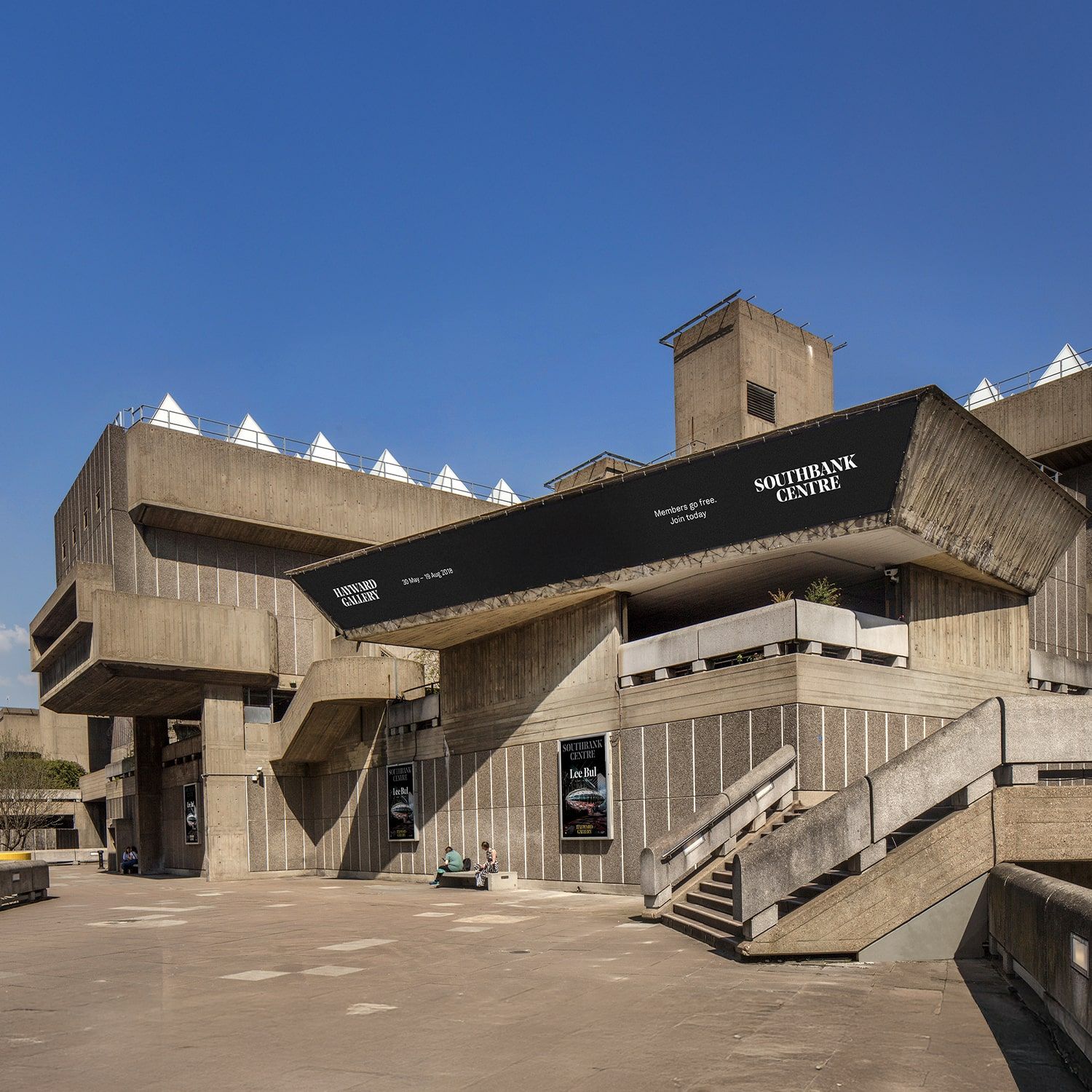 A large Brutalist concrete building with angular shapes and ramps, featuring a prominent black sign reading Southbank Centre. Few people sit on benches in the open plaza under a clear blue sky.