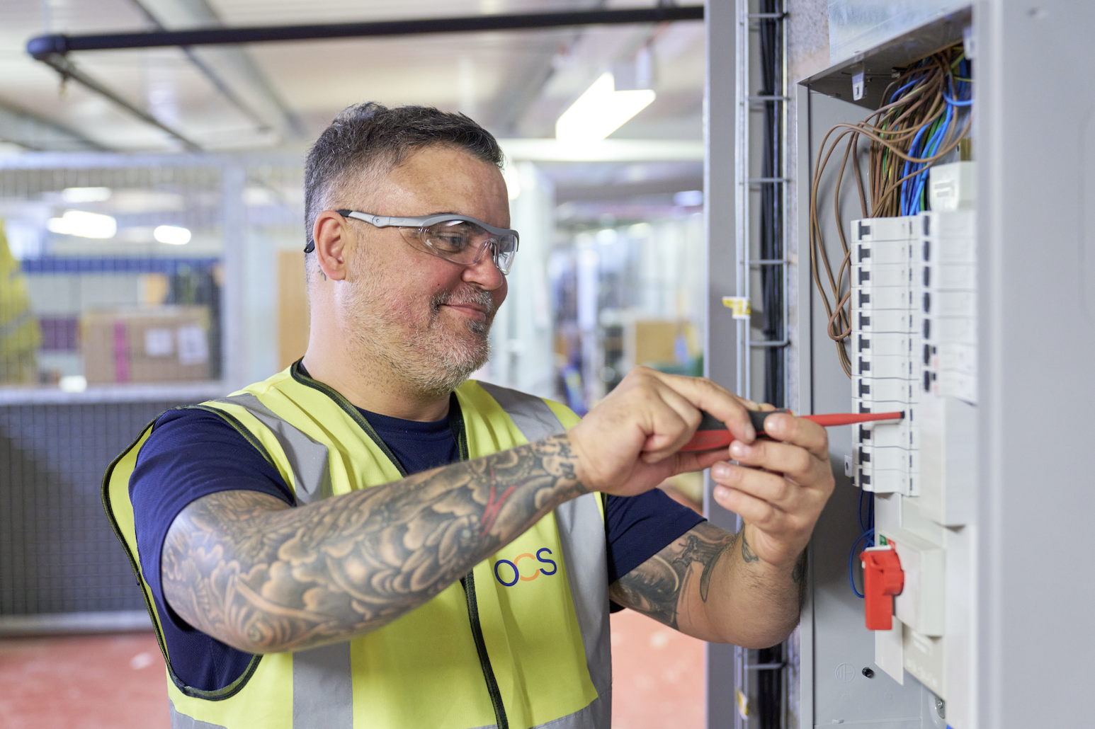 A man wearing safety glasses and a high-visibility vest uses a screwdriver to work on an electrical panel. He has visible tattoos on his arm and is focused on his task in an industrial setting.