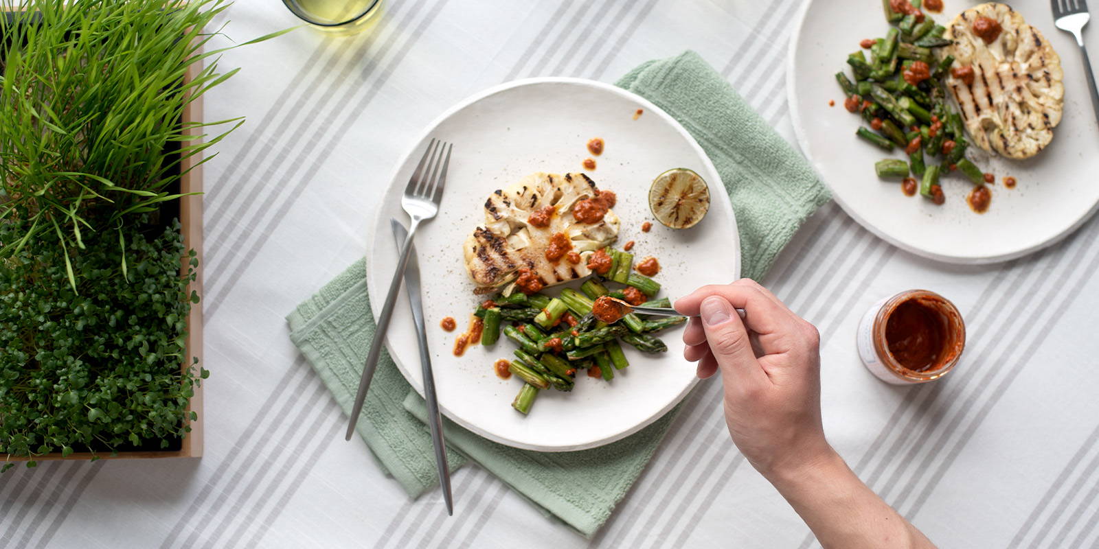 A white plate with a slice of grilled cauliflower and bite-sized grilled asparagus, being garnished with harissa. On the side of the plate sits a grilled lime half, and in te upper right is a second plate of grilled cauliflower and asparagus, already garnished with harissa and lime.