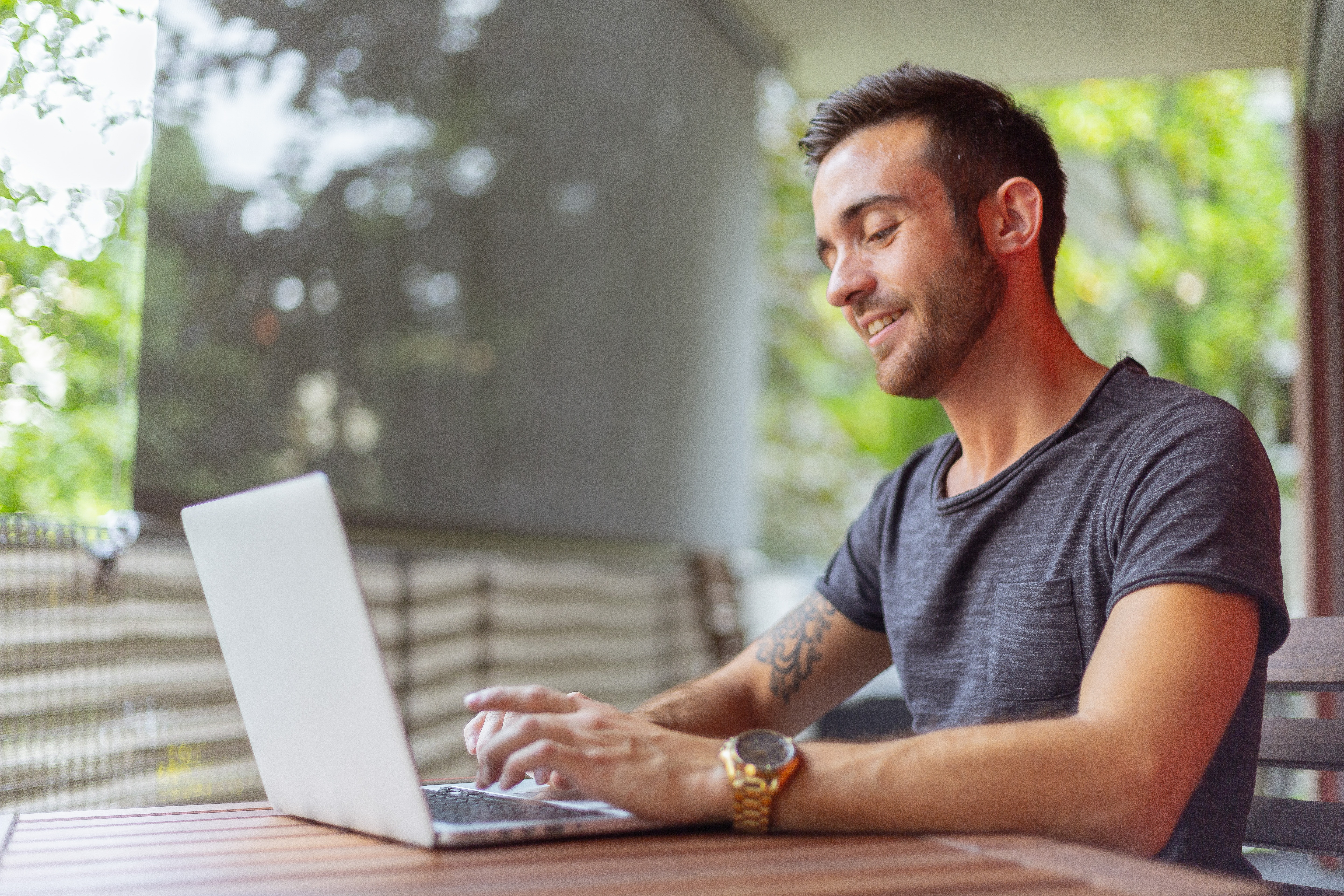 Image of an young white man with a beard tattoo, sitting in an open area typing on his laptop and smiling.