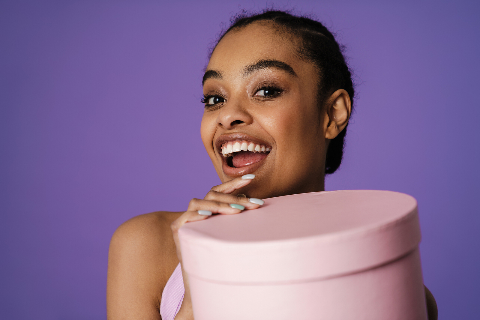 An attractive young black woman with her hair back and a pink shirt holding a circular pink box with a big smile on her face ready to open the box.
