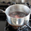 A stainless steel saucepan containing a dark liquid is simmering on a gas stove burner, with steam rising from the surface.