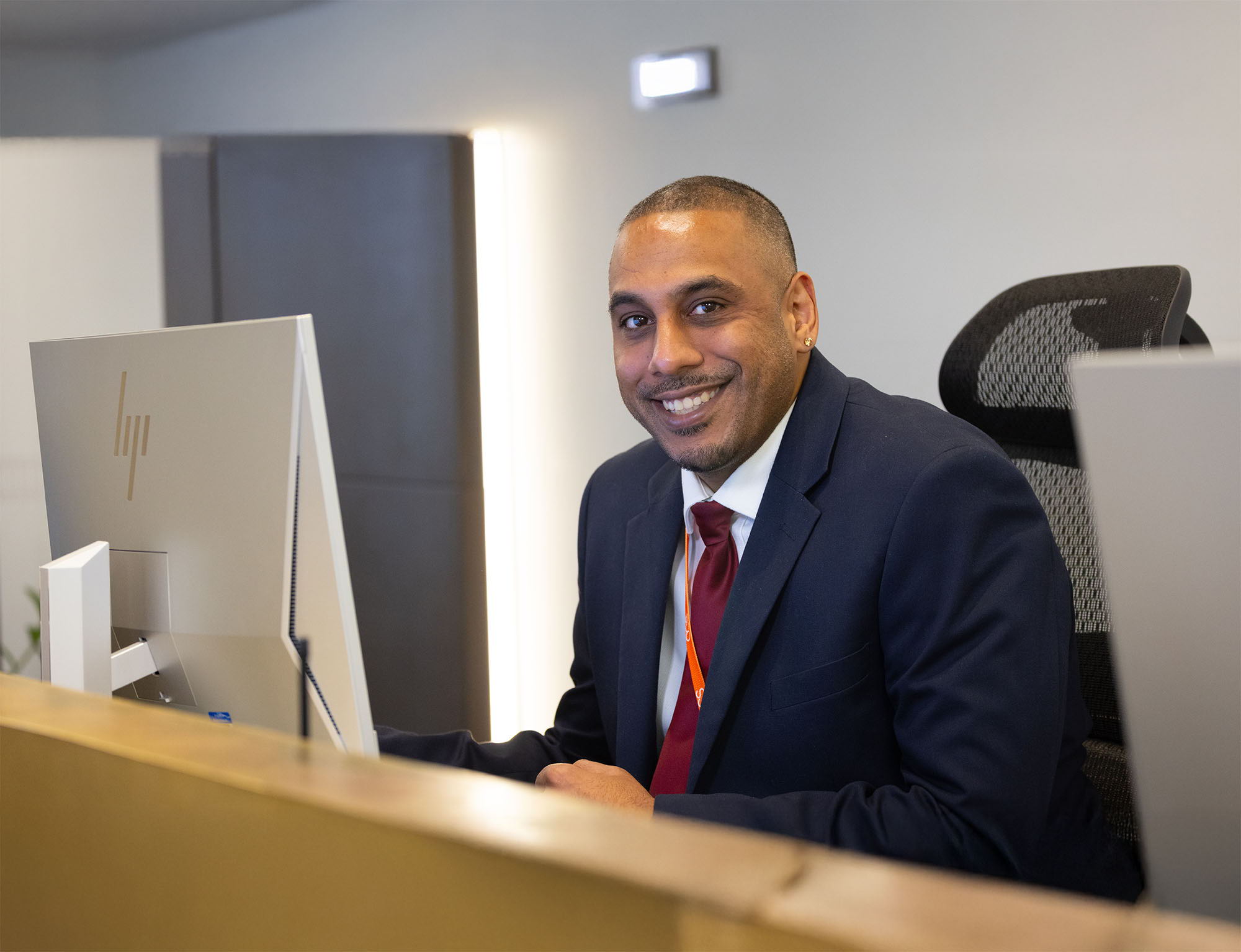 A man in a suit and red tie sits at a desk in an office, smiling at the camera while working on a computer.
