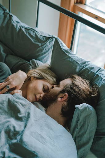 A couple snuggled up in bed while the man gives the woman a kiss on the forehead - Photo by Toa Heftiba on Unsplash