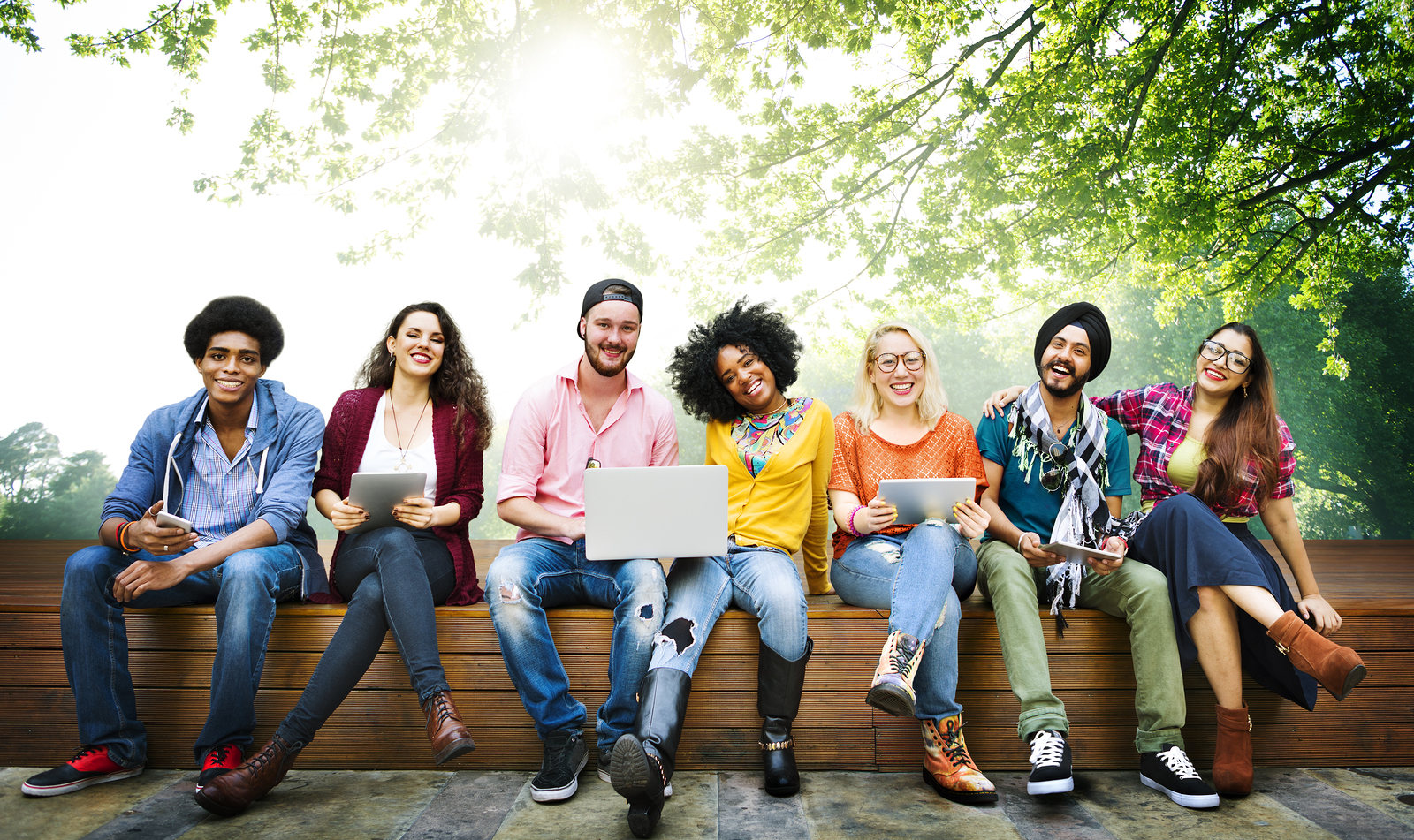 A large grou pf multi ethnic friends sit together on a bench smiling to the camera with laptops and tablets in front of them.