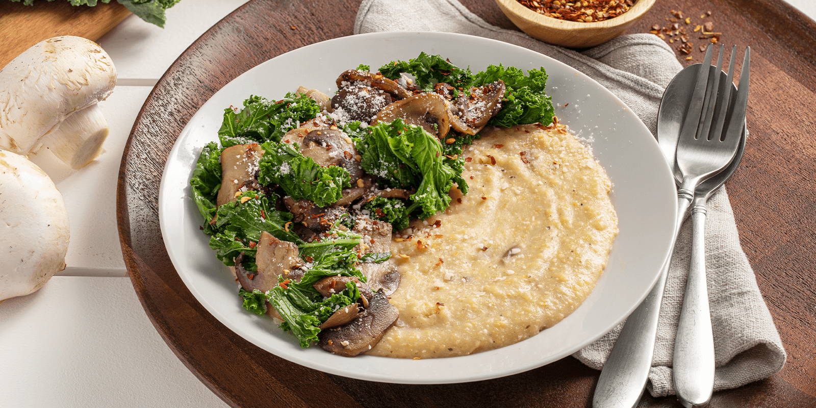 The prepared ZENB Creamy Mushroom Polenta with Kale & Garlic in a shallow white dish atop a wooden cutting board with silverware on a gray napkin on the side. 
