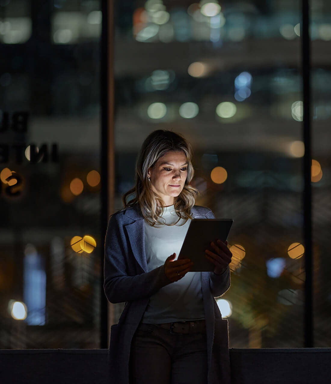 A woman stands indoors at night, illuminated by the light from a tablet she is holding, with blurred city lights visible through large windows in the background.