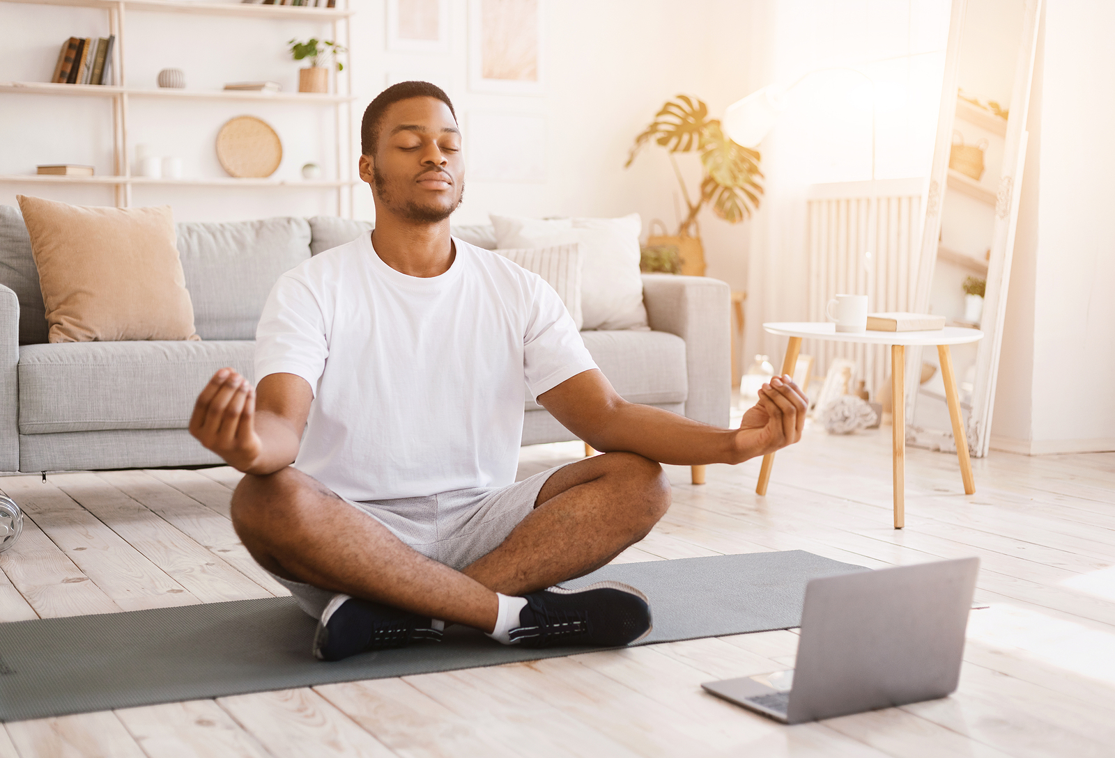 A black man meditating on the floor with a computer nearby, he is calm and smiling.