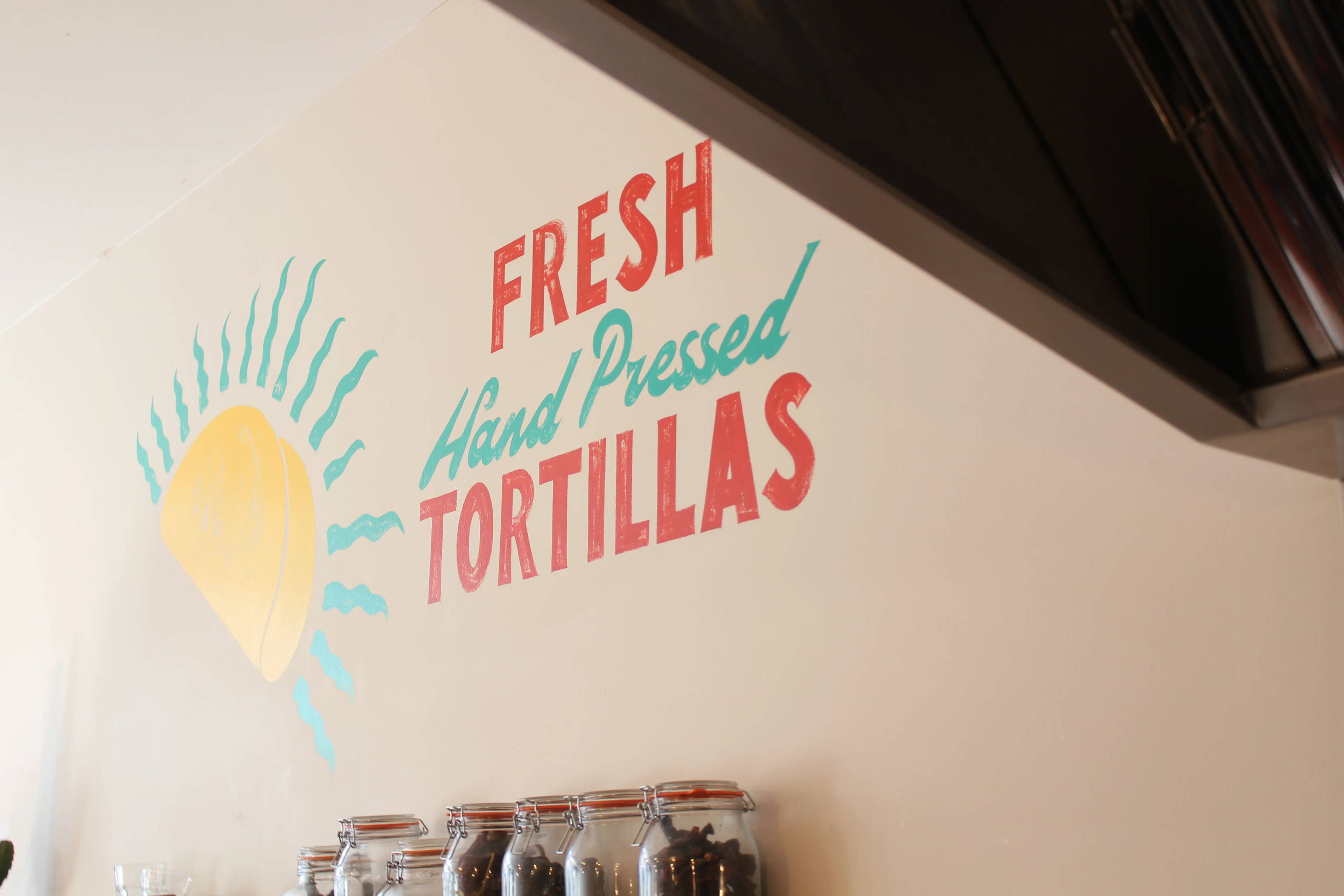 A hand-painted red and green sign on a cream wall that read "Fresh hand pressed tortillas"