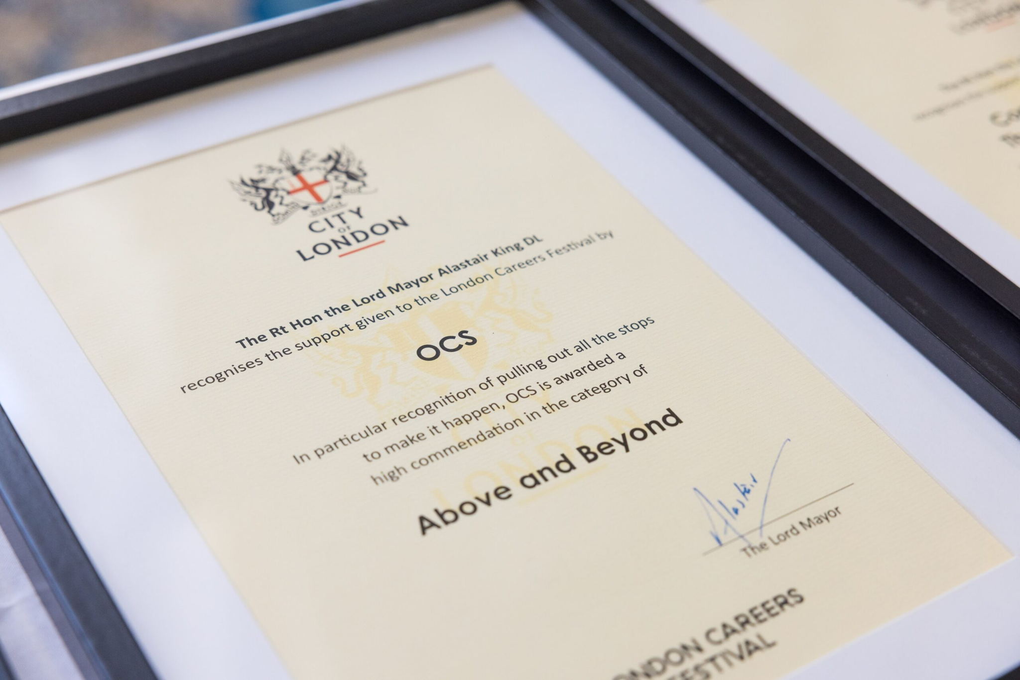 A framed certificate from the City of London awarded to OCS, recognizing outstanding support at the London Careers Festival, with a signature and the title Above and Beyond visible on the document.
