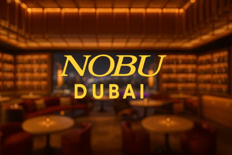 Warmly lit interior of Nobu Dubai, featuring blurred tables, plush seating, intricate shelving, and a city skyline view through a large window, with the restaurant name overlaid.