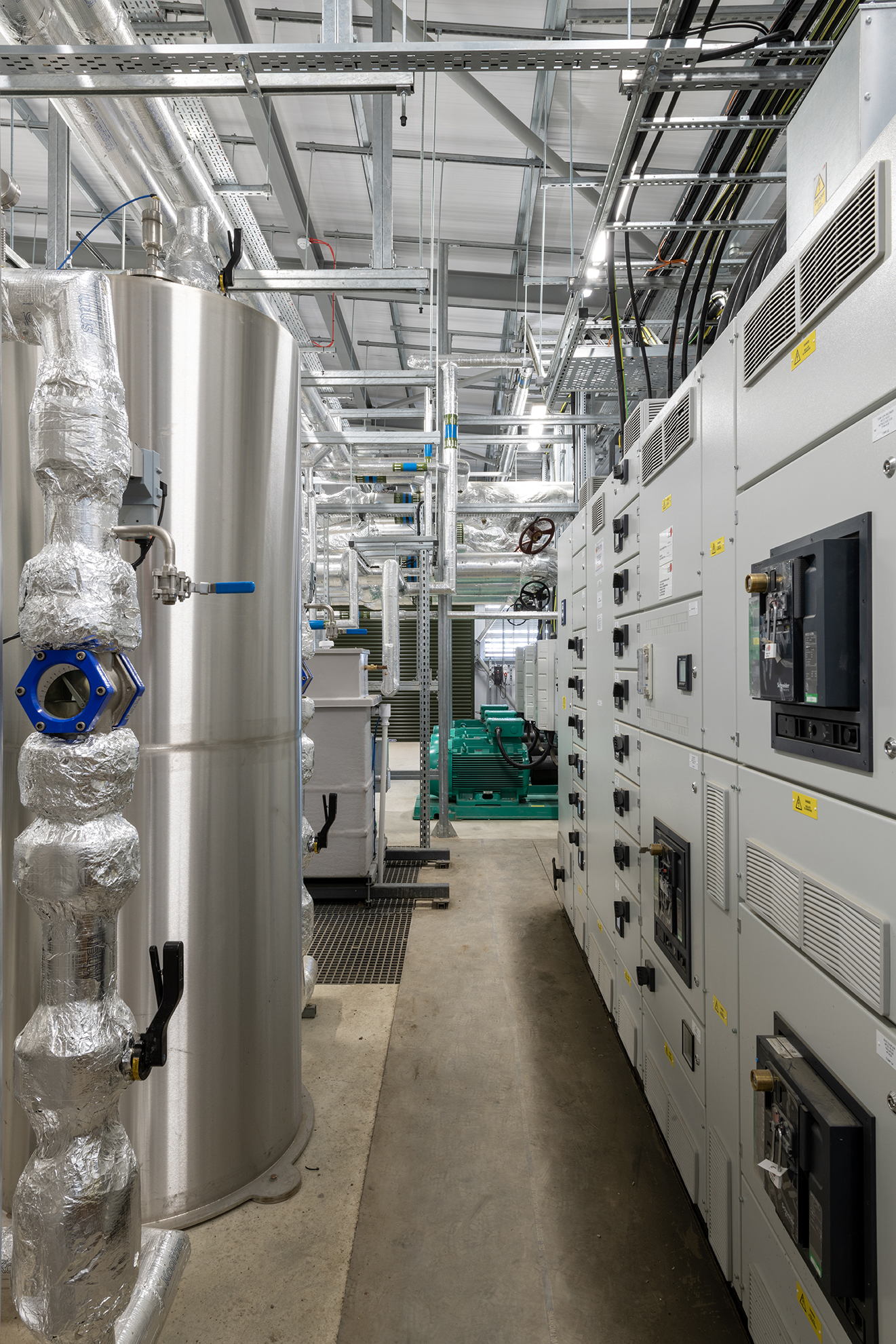 A modern industrial facility interior showing large metal pipes wrapped in insulation on the left, electrical control panels on the right, and exposed ceiling with cables and ducts above.