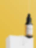 A sleek bottle of face serum with a dropper, displayed on a white pedestal against a vibrant yellow background.