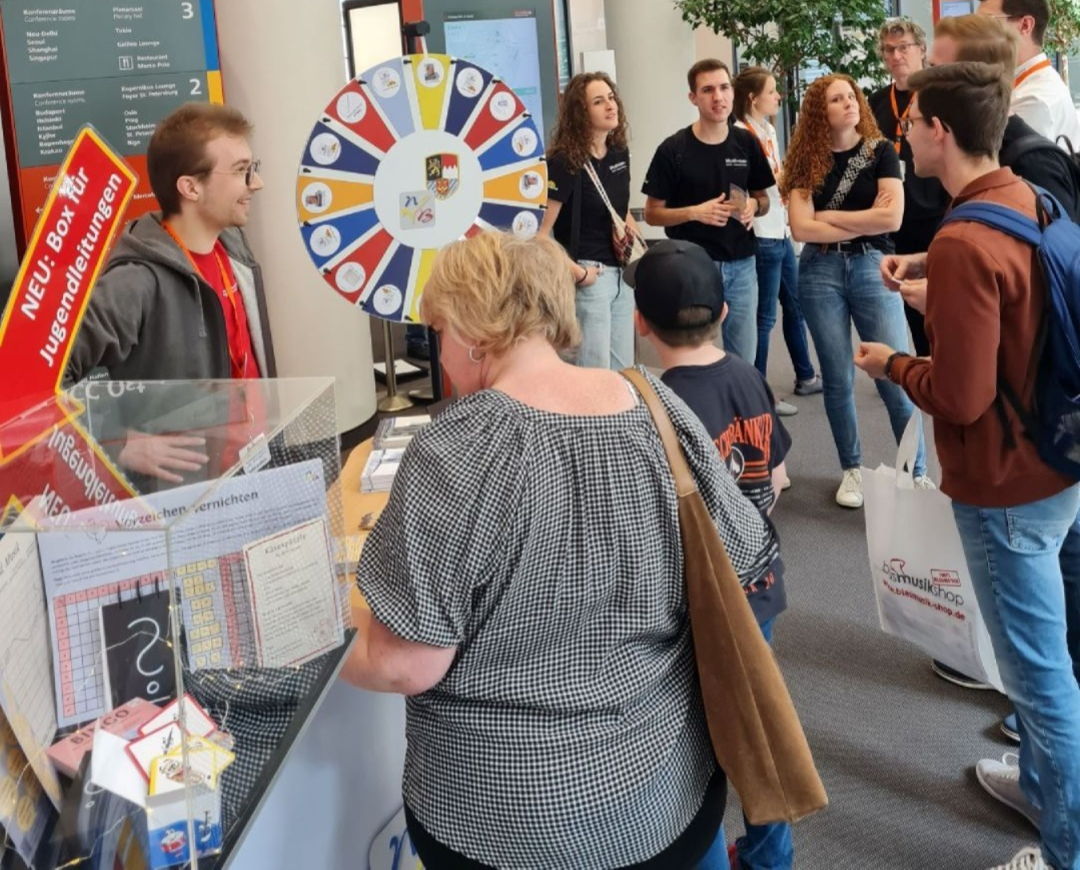 An einem Messe-Stand mit einem Glücksrad stehen viele Menschen und unterhalten sich. 