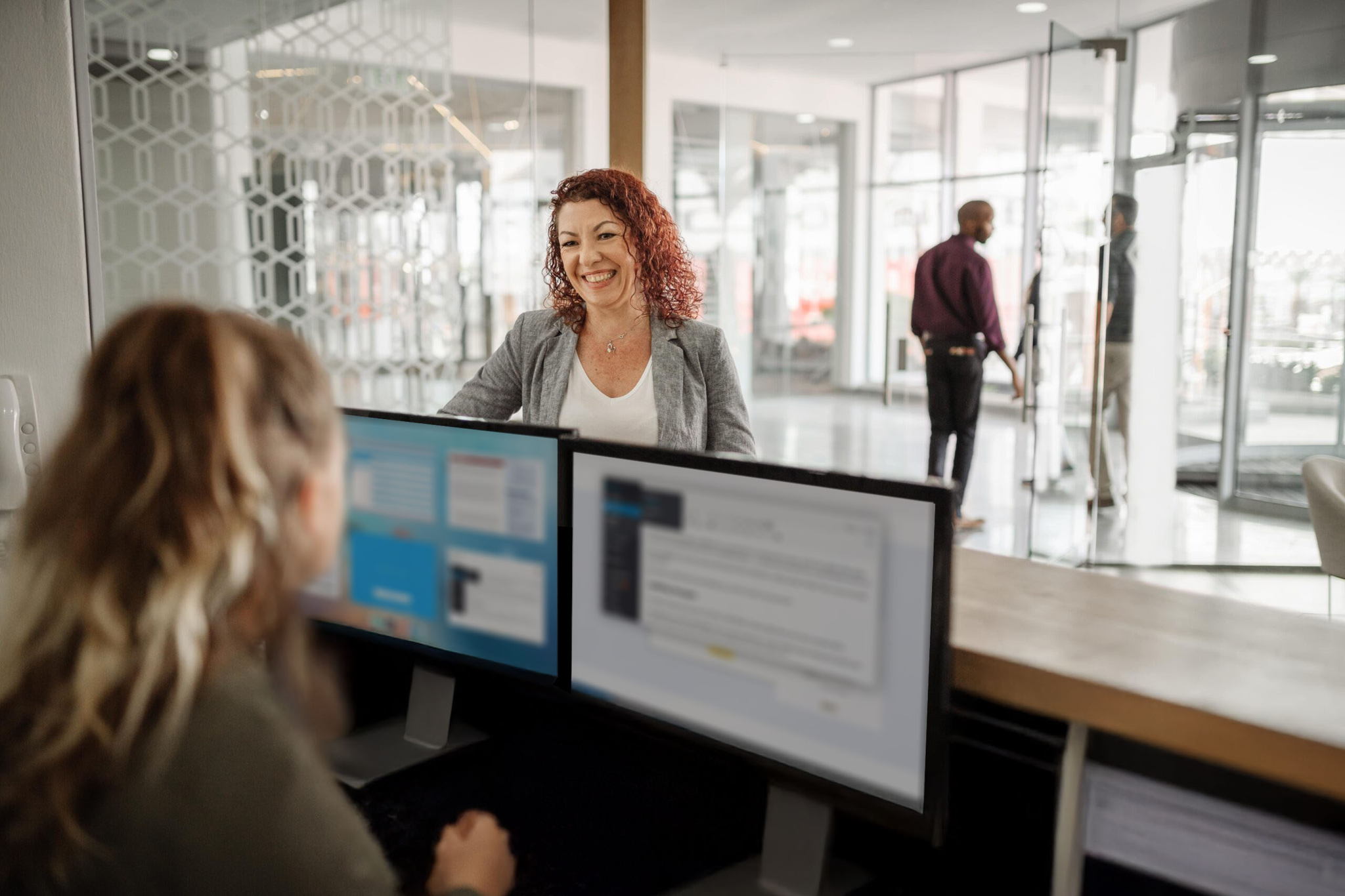 A woman with curly hair is smiling at a reception desk where another person is seated, working on dual monitors. In the background, two people are walking in a modern office lobby with glass walls.