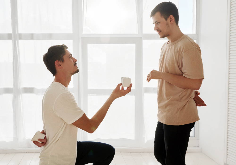 A kneeling man offers another man a small box containing an engagement ring.