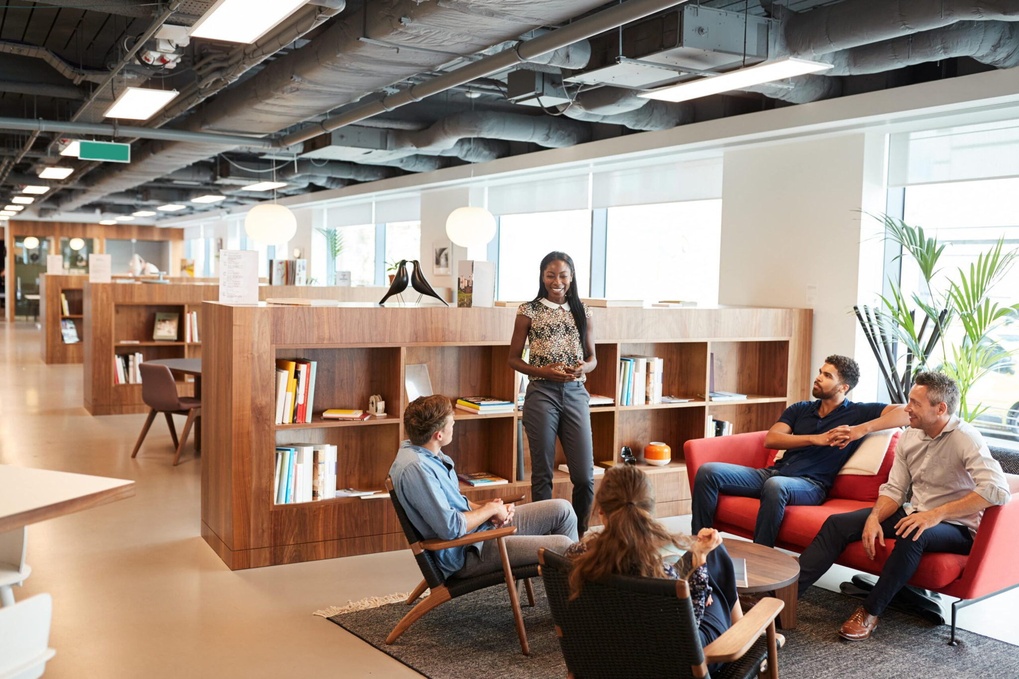 A woman stands and speaks to four seated colleagues in a modern office, designed through our workplace services, with wooden bookshelves and sofas. Large windows let in natural light. The group is gathered around a small table on a rug, creating a collaborative atmosphere.