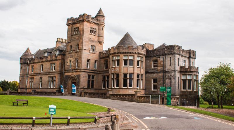 A large, historic stone castle with towers, arched windows, and a circular turret sits beside a curved road, surrounded by green grass and trees under a cloudy sky.