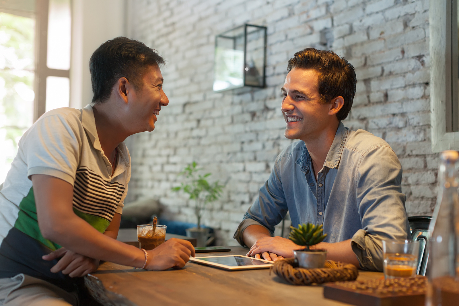 Two men of different ethnicities sitting together at a cafe, smiling to eachother