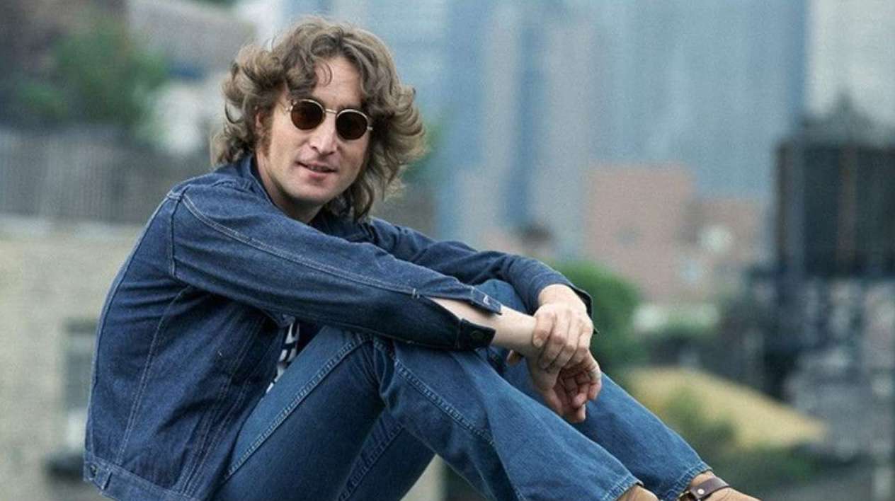 John Lennon with his hands on his knees sitting looking at the camera wearing sunglasses.