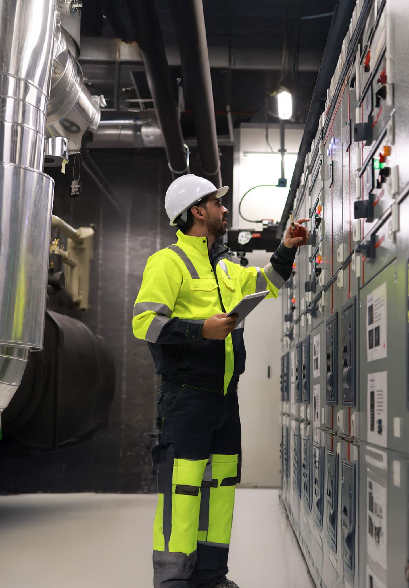 A worker wearing a high-visibility jacket, helmet, and holding a clipboard is adjusting controls on a large panel in an industrial setting. Pipes and machinery are visible in the background.