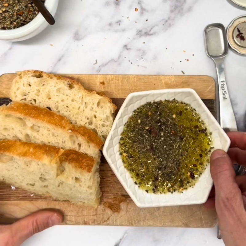 A wooden cutting board with slices of bread on it next to a white dish of oil and spices
