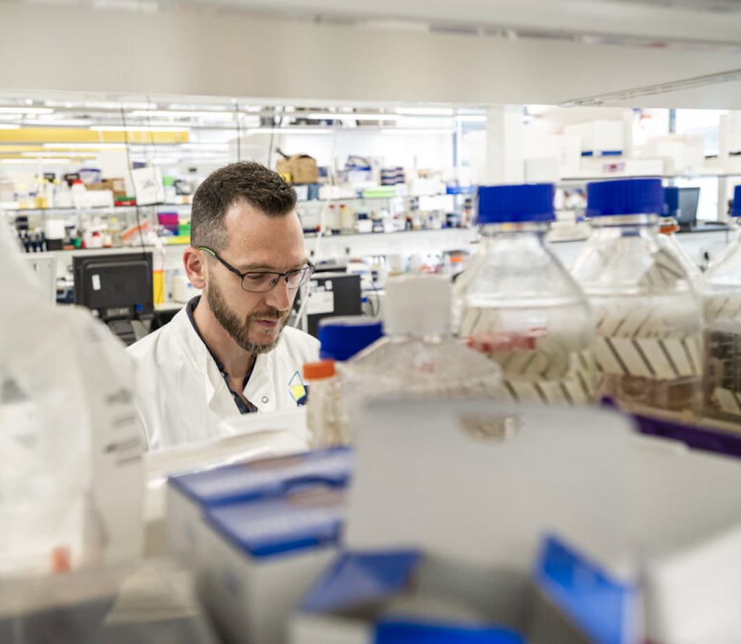 A scientist in a white lab coat works in a bright laboratory, surrounded by various bottles, containers, and lab equipment on the workbench.