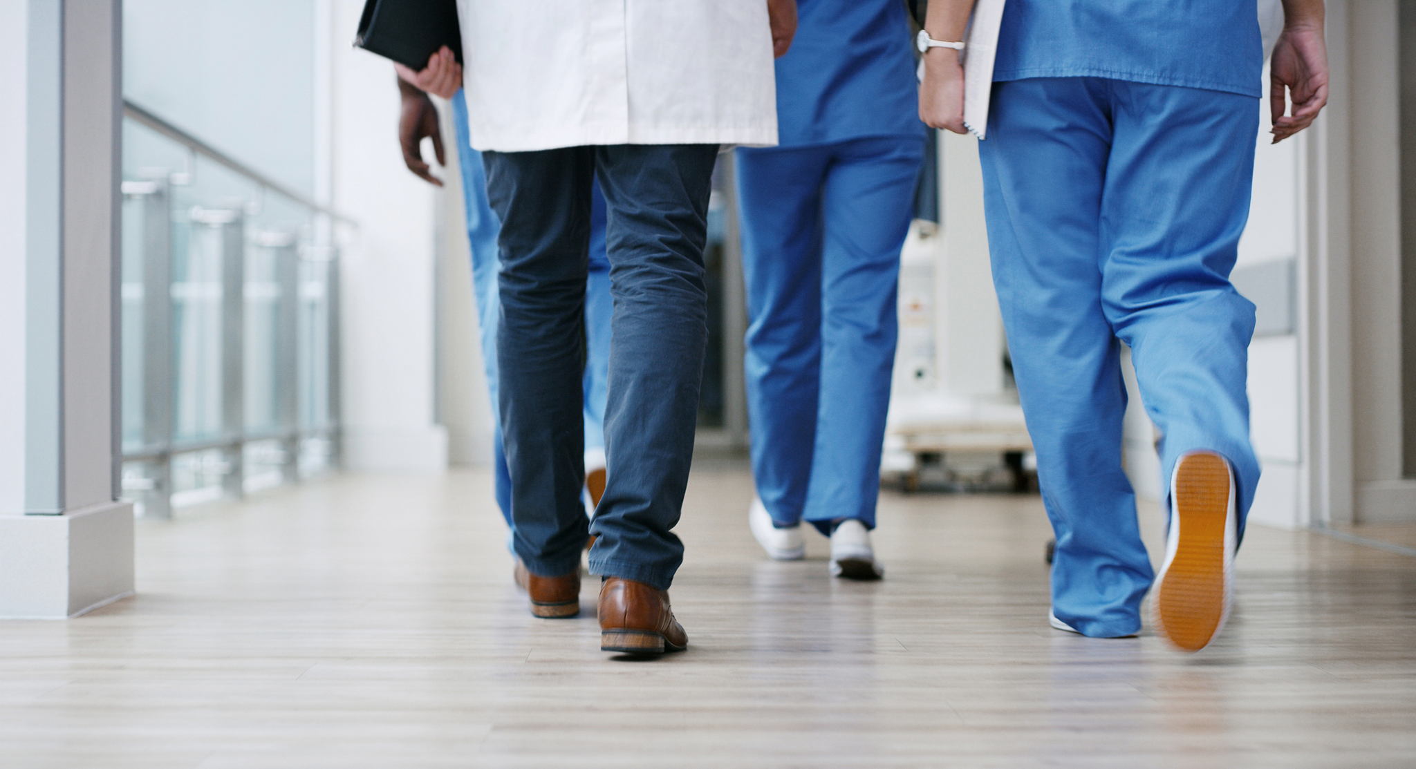 Three healthcare professionals, seen from behind and wearing scrubs and lab coats, walk down a hospital corridor with bright lighting and a wooden floor.