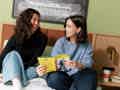 Zwei Frauen sitzen lächelnd auf einem Hotelbett, halten ein Playbill-Magazin in der Hand und haben Speisen und Getränke auf dem Nachttisch.