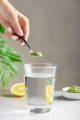 lady's hand holding a small spoon of wheat grass powder over a clear glass of water, with lemon in the background