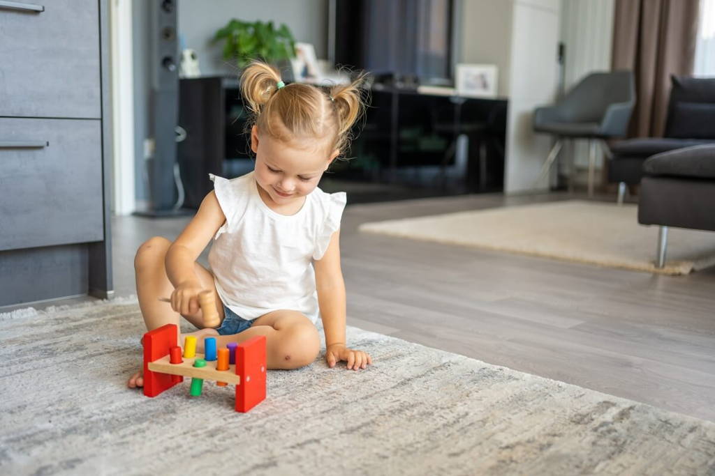 Little girl sitting on a rug and playing with a wooden hammering toy.