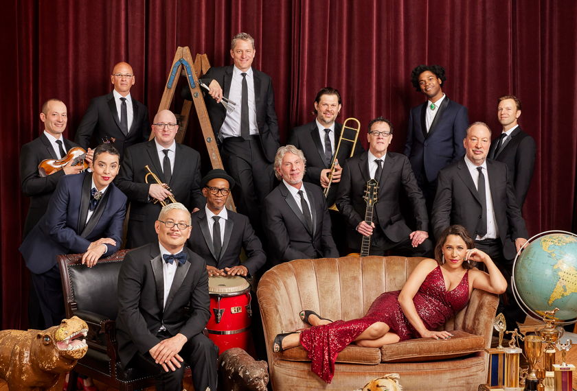 Members of Pink Martini are dressed in formal attire in front of a red, velvet curtain. 