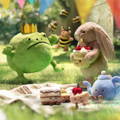 Jellycat sad looking frog and bashful bunny holding a birthday cake at a picnic with other Jellycat food items and teapot, with Jellycat bees buzzing around.