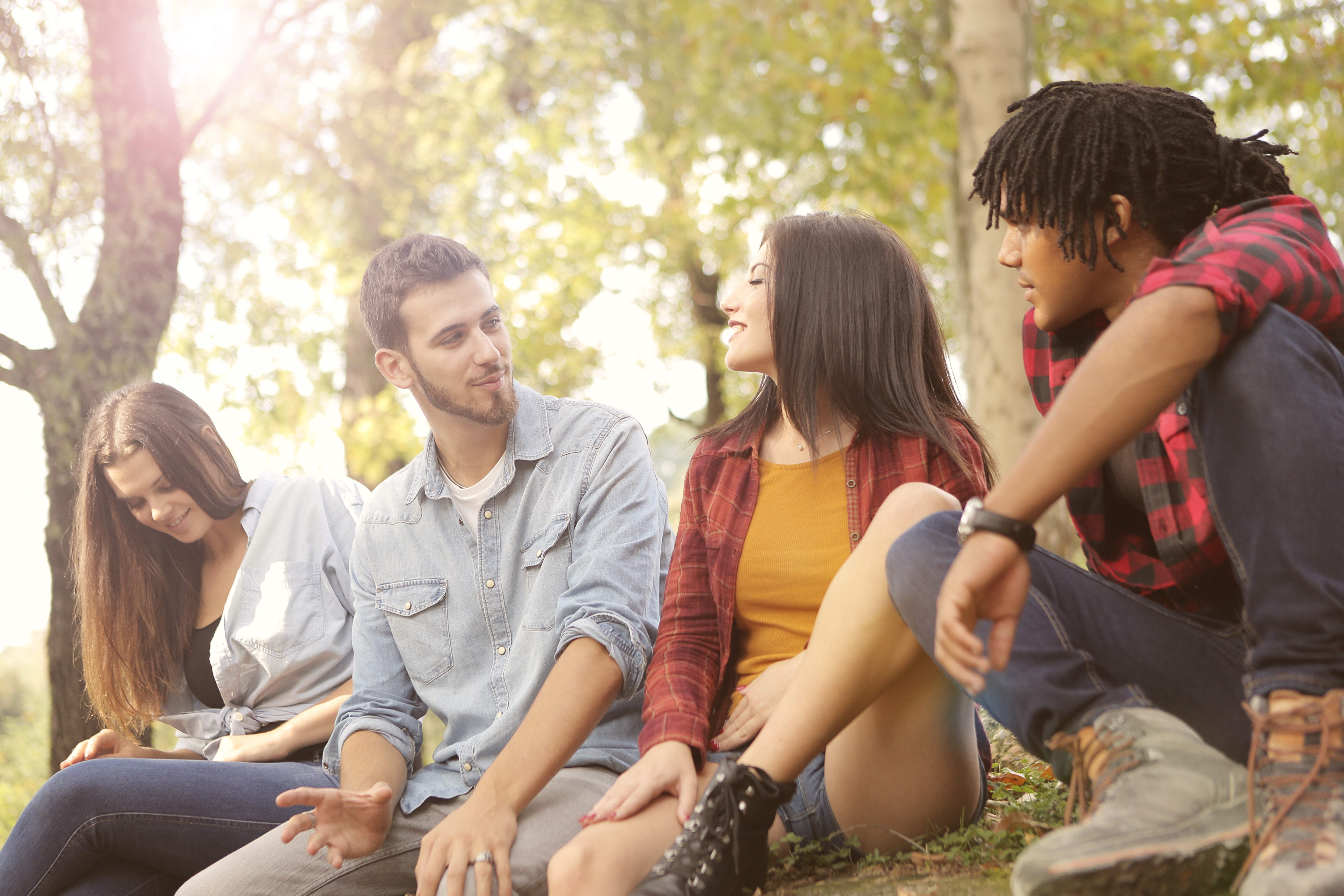 Image of a group of 4 multi ethnic and attractive men and women friends, talking to eacth other smiling sitting in a park.