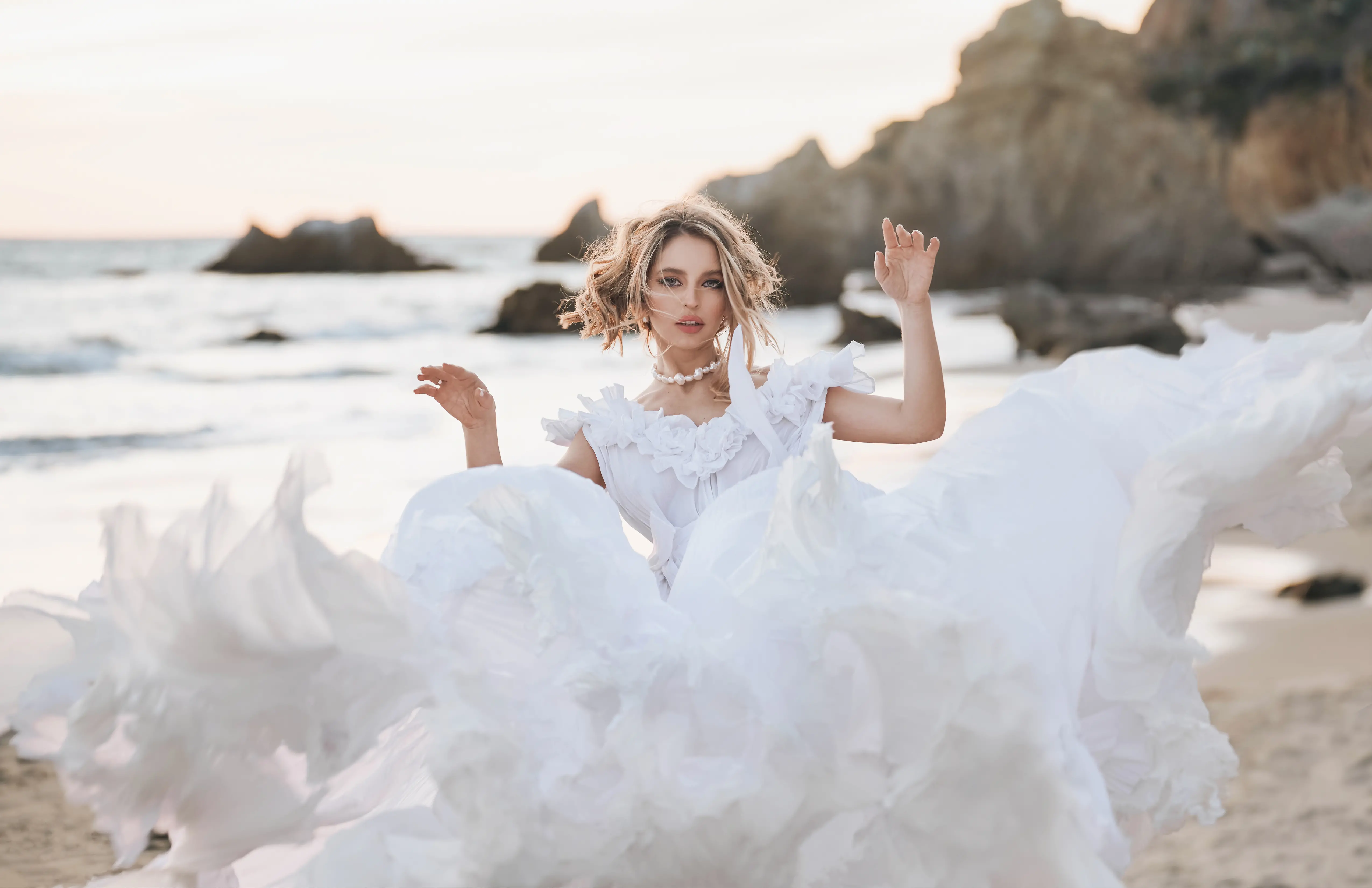 Une femme debout sur la plage vêtue d'une robe de mariée blanche, avec les vagues de l'océan en arrière-plan. La robe, agitée par le vent, ajoute une touche d'effervescence au cadre côtier.