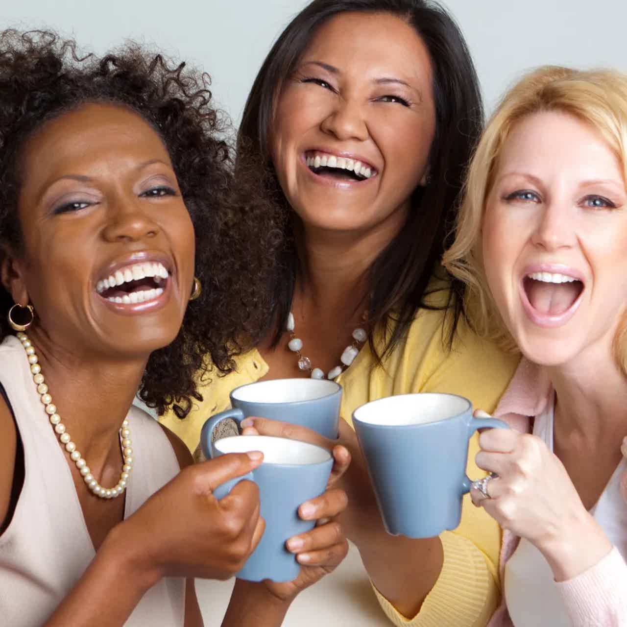 Three women smiling and cheering with mugs, toasting drinks. Bright, joyful group portrait.
