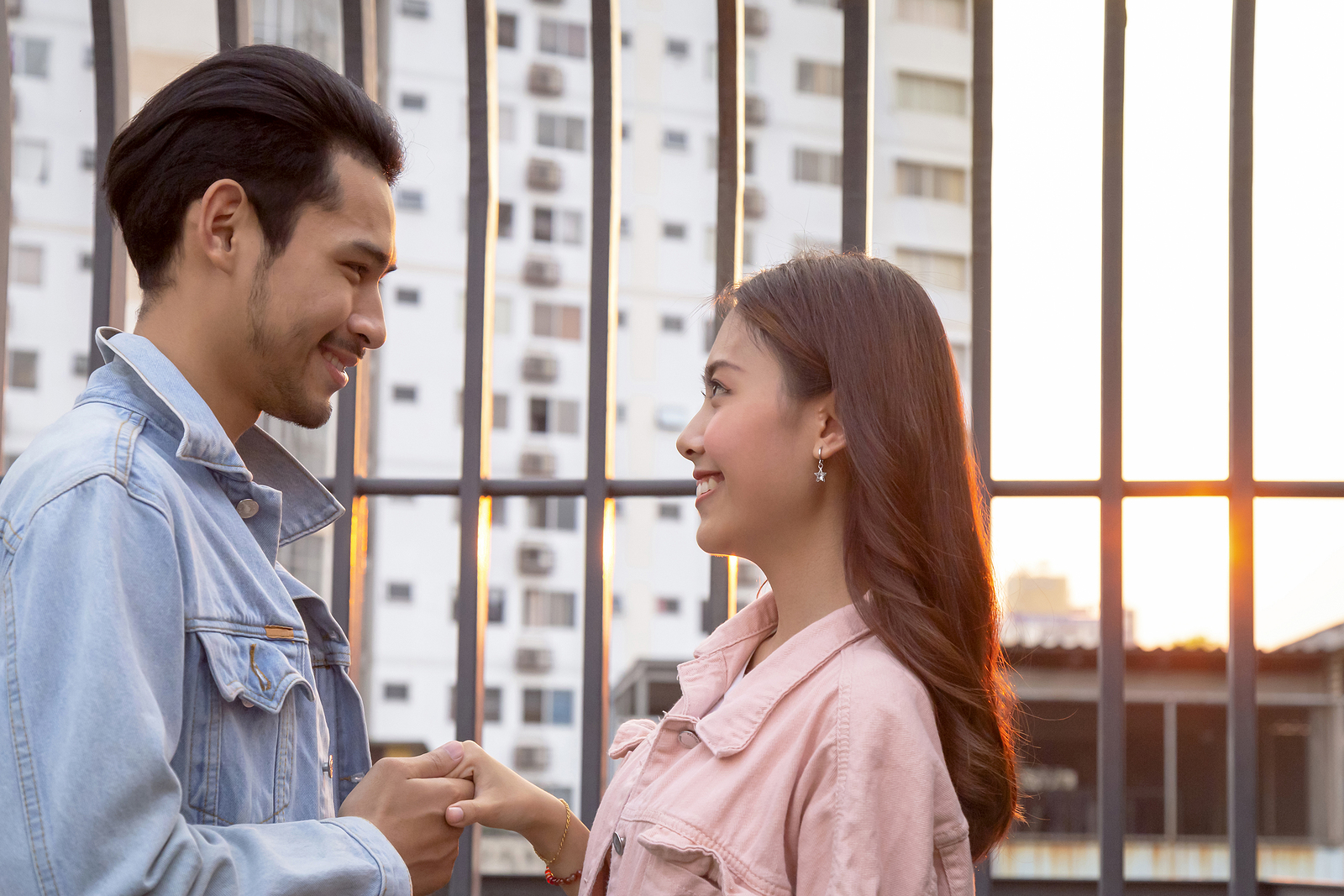 A young asian man and woman stand facing eachother. They are holding onto a hand and smiling.