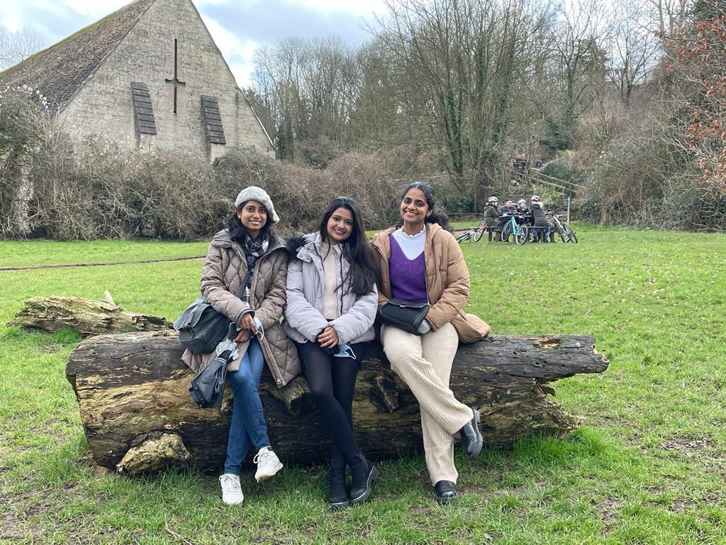 Gayathri (right) on a day out with her flatmates. They are sitting on a log in a green field.