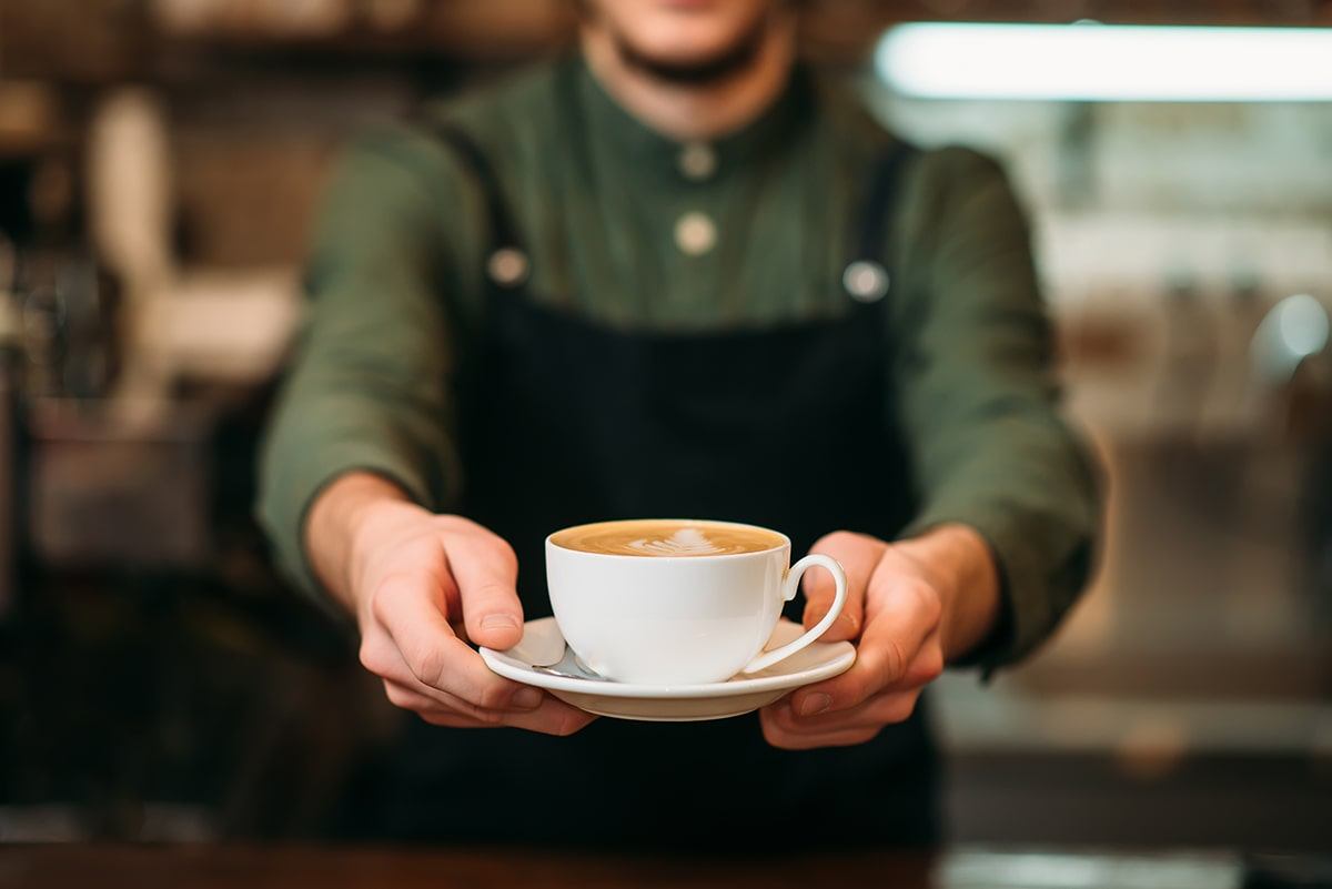 A barista wearing a dark apron and green shirt presents a cup of cappuccino with latte art on top, holding the white cup and saucer with both hands toward the camera.
