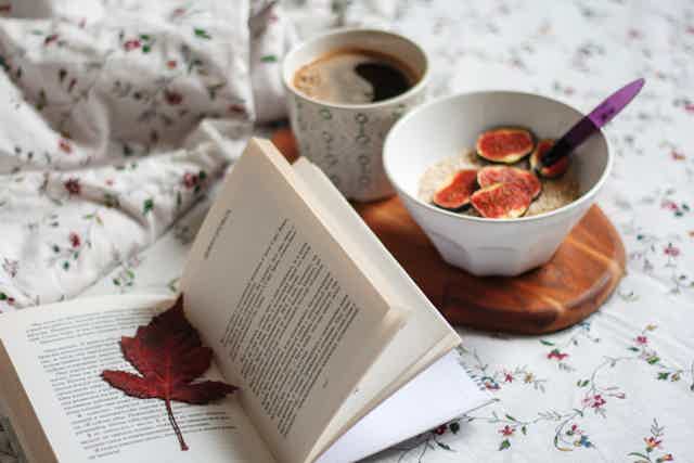 Open book on a blanket with a fallen leaf, a cup of coffee, and a bowl of figs on a wooden tray.
