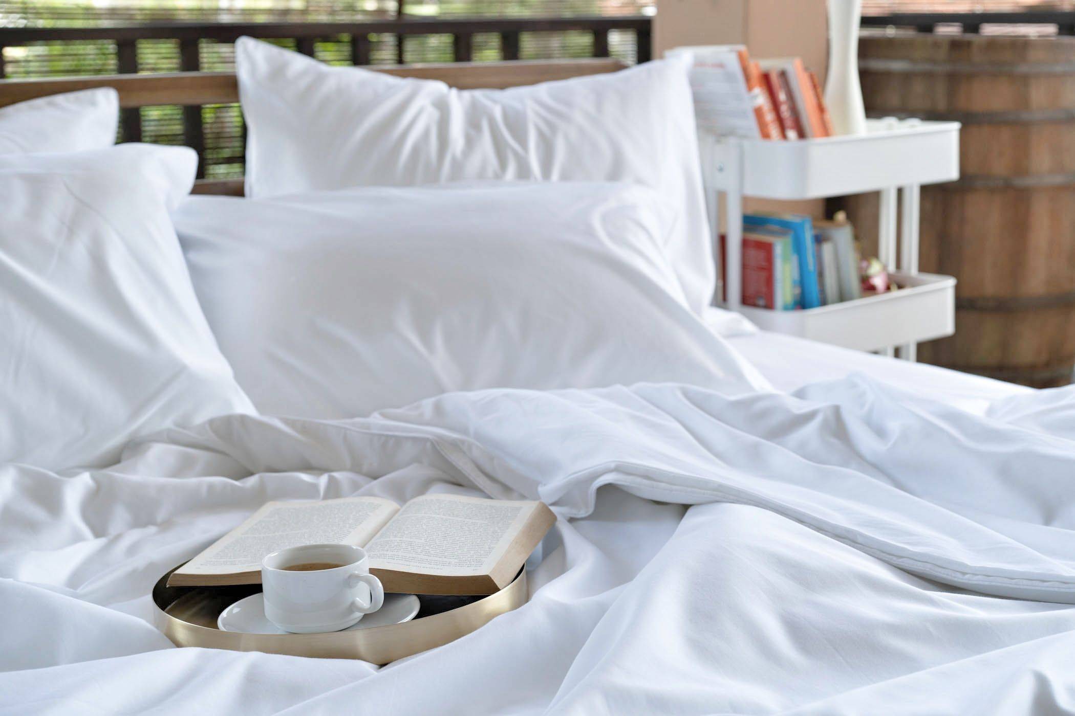 Featuring Weavve's Cloud White Cotton Deluxe Set with 1 fitted sheet, 1 duvet cover, 4 pillow cases and 2 bolster cases, with a tray with a coffee mug and book placed on top of it