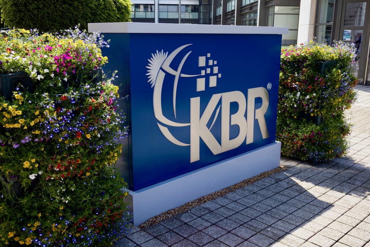 A large blue sign with the KBR logo and stylized globe design stands outdoors, surrounded by colorful flowering plants and a paved walkway.